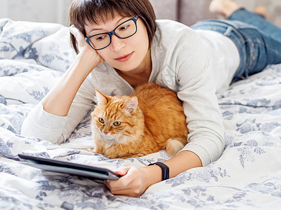 可爱的姜猫和戴眼镜的女人躺在床上 女人拿着平板电脑摸着她的毛绒宠物 早安时间就寝屏幕猫咪动物药片技术女性女士哺乳动物图片