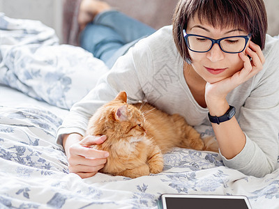 可爱的姜猫和戴眼镜的女人躺在床上 女人在看平板电脑 摸着她毛茸茸的宠物 早安图片