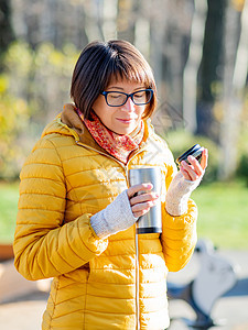 穿着亮黄色夹克的快乐笑容的女人拿着热水杯 在寒冷的秋天喝热茶或其他饮料旅行公园晴天女孩阳光木头游客烧瓶情感森林图片