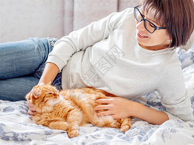 可爱的姜猫和戴眼镜的女人躺在床上 女人在摸她的毛绒宠物 早安哺乳动物卧室毯子情感猫科动物就寝投标时间女士动物图片