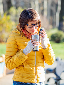 穿着亮黄色夹克的快乐笑容的女人拿着热水杯 在寒冷的秋天喝热茶或其他饮料幸福女士女孩森林女性金属情感游客木头咖啡图片