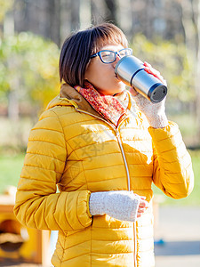 穿着亮黄色夹克的快乐笑容的女人拿着热水杯 在寒冷的秋天喝热茶或其他饮料旅行游客眼镜情感阳光女孩女性保温瓶晴天旅游图片