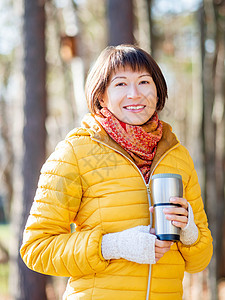 穿着亮黄色夹克的快乐笑容的女人拿着热水杯 在寒冷的秋天喝热茶或其他饮料女性烧瓶情感女孩旅行森林女士咖啡木头旅游图片