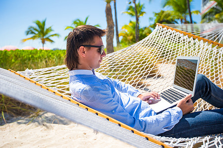在沙滩度假期间在吊床上用笔记本电脑工作的年轻人支撑上网互联网海岸人士热带记事本商务太阳镜游客图片
