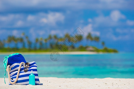 条纹袋 蓝毛巾 太阳墨镜 防晒霜瓶和泳衣 背景包括绿绿绿水和绿棕榈树图片