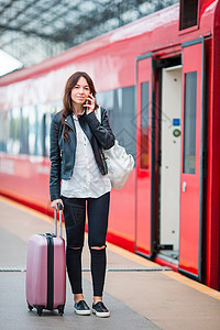 携带行李的年轻女子在火车站用手机交谈 她旅行时乘快车等候客服旅游者 笑声旅游速度城市女孩铁路电话车皮火车木板乘客图片