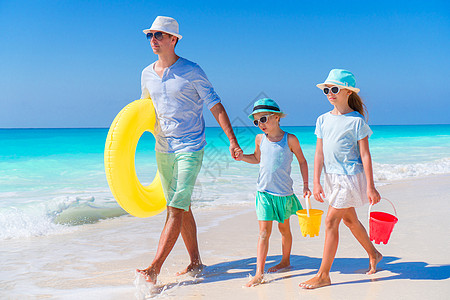 父亲和孩子的家庭在卡里比恩岛的白色热带海滩上行走 并配有海滩玩具图片