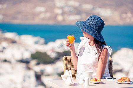 在Mykonos镇景色惊人的咖啡厅里喝咖啡的年轻女子奢华饮食阳台早餐酒店假期咖啡店桌子水果城市图片