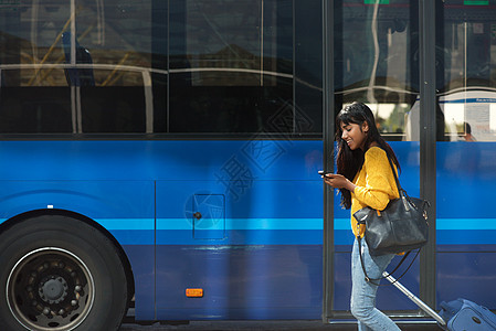 带着手提箱和手机在公共汽车车站行走的微笑着笑着的年轻印度妇女图片
