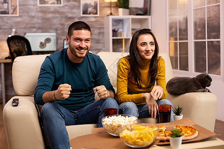 快乐的情侣为他们最喜欢的团队欢呼 同时在客厅看电视房子成人电视夫妻午餐公寓男性扇子沙发流行音乐图片