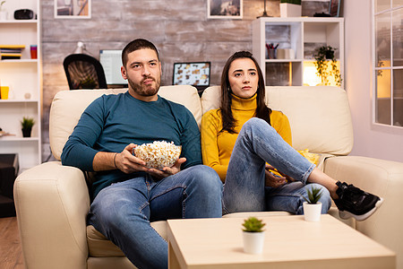 在家吃流行玉米和看电视的一对英俊夫妇爆米花食物休息男人家庭电影乐趣电视长椅沙发图片