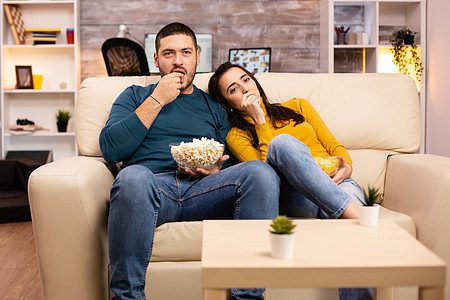 在家吃流行玉米和看电视的一对英俊夫妇微笑男朋友休息房子男性沙发喜悦房间家庭电影图片