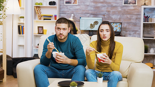 坐在沙发上坐着的一对夫妇 用筷子吃面女士电视美食食物长椅命令闲暇电影夫妻微笑背景图片