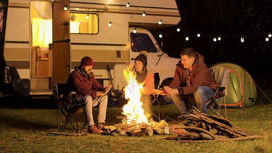 朋友们在寒冷的夜晚 在营火中暖热手团体旅游火树林帐篷森林火焰露营者胡子营火椅子图片