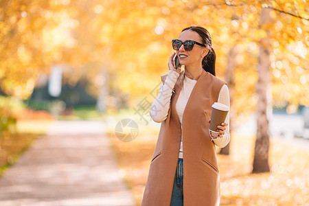 秋瀑概念  美丽的女人在秋公园喝咖啡 在秋天的树叶下饮料杯子手机女士快乐季节外套木头叶子女孩图片