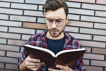 穿着眼镜的年轻嬉皮士 在读关于砖墙背景的书或笔记本衬衫成人学习学生教育知识文学笔记大学男性图片
