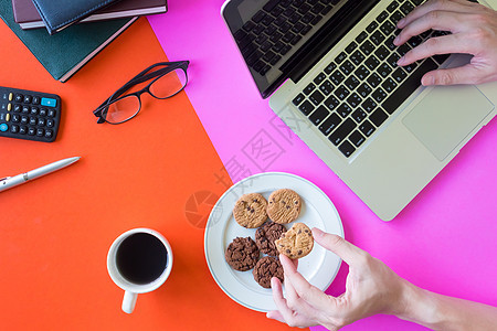 男性手拿着饼干 使用笔记本电脑 咖啡杯和办公室用品 背景丰富多彩图片