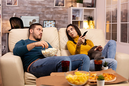 美丽的年轻夫妇在看电视和吃快餐外卖电视午餐手表食物享受电影房间苏打流行音乐爆米花图片