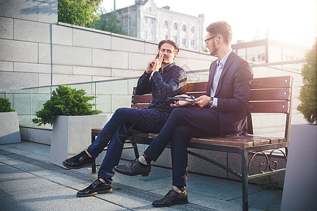 两位坐在街上长椅上的商务人士 其中一人的电话太忙了技术成人成功男人工人药片公园讲话团队职业图片