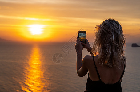 女人手里拿着手机在日落时分 年轻的卷发女人在海上美丽的夕阳中用手机拍照 在假期 旅行期间在智能手机上拍照细胞电话海洋天空女士橙子图片