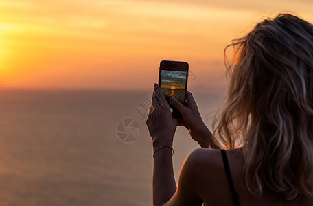 女人手里拿着手机在日落时分 年轻的卷发女人在海上美丽的夕阳中用手机拍照 在假期 旅行期间在智能手机上拍照相机技术电话旅游屏幕风景图片