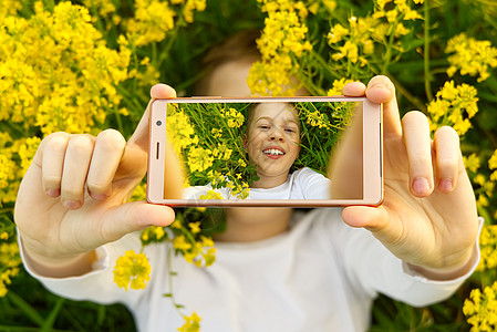 快乐的微笑男孩在草地上用智能手机制作自画像 的小男孩躺在开着黄色花朵的绿草地上的智能手机上自拍公园微笑电话喜悦母亲照片女士女孩假图片