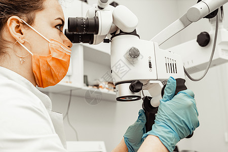 年轻的女牙医通过牙科诊所的专业显微镜进行观察 戴一次性医用口罩和帽子的医生 牙科先进设备成人操作从业者助手保健实验室程序医学口腔图片