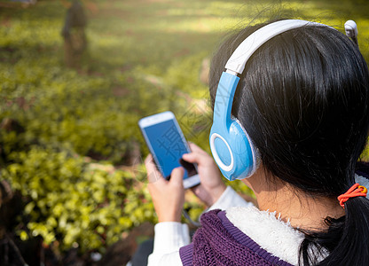 亚洲年轻女孩在花园里用耳机听音乐 亚洲年轻女孩在花园里用耳机听音乐 技术和放松的概念背景图片