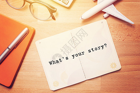你的故事是什么? 带空白页文字的笔记本 在木形背景上印有眼镜和飞机模型图片