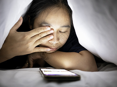 使用智能手机的亚洲女性肖像躺在床上的白色毯子下 眼睛疼痛 她的脸被明亮的监视器照亮 休息时间互联网闲暇技术阅读就寝女士电话房间卧图片