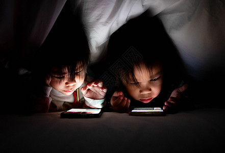床上毯子下的智能手机上 亚洲小女孩和妹妹的肖像看起来很卡通 明亮的监视器照亮了孩子的脸 保健概念女士游戏房间乐趣技术卡通片眼睛家图片