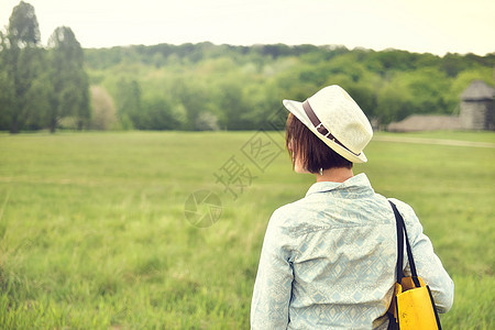 身着衬衫 草帽和袋子的黑发女人在夏季田间行走时背影图片