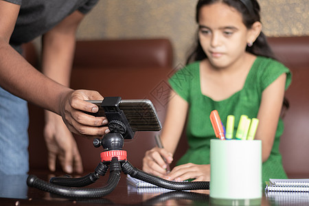 父亲通过调整手机来帮助女儿学习在线课程或电子学习的课程-父母在虚拟课堂 家庭教育 远程学习中支持孩子的角色概念图片