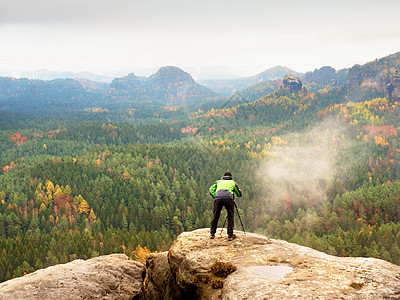带相机的徒步旅行者在三脚架上从岩石山顶拍照 边缘照片风景的单独摄影师石头夹克装备条件登山服远足者假期首脑橙雾单反背景图片