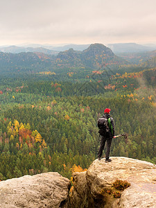 高高的徒步男子带红帽 手握电杆 在岩石上摆着大背包架导游幸福首脑棒球帽冒充顶峰背包悬崖爬坡登山者图片