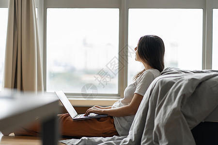 美丽的年轻黑发女孩在笔记本电脑上工作 坐在靠近全景窗户的床边的地板上 时尚现代的内饰 舒适的工作场所 在互联网上购物学生网络商业图片