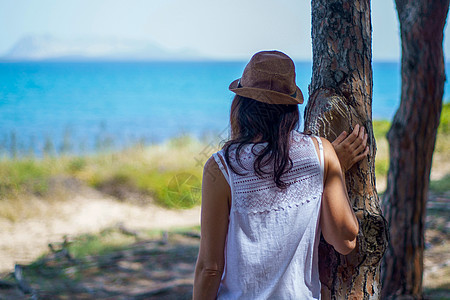 从后面看到长头发和帽子的年轻女性 俯视着在松树林中一棵树上倾斜的海安详海洋沉思太阳成人寂寞日落思维享受女士图片