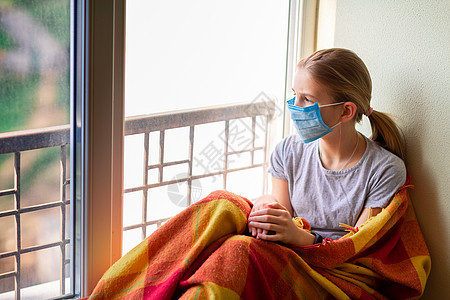戴面罩的可怜小女孩坐在家里被隔离的窗户上 Corona病毒检疫概念图片