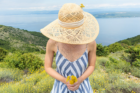 穿着条纹夏装和草帽的年轻女子站在野花盛开的地方 在克罗地亚亚得里亚海沿岸自然的美丽大自然中捧着一束黄花图片