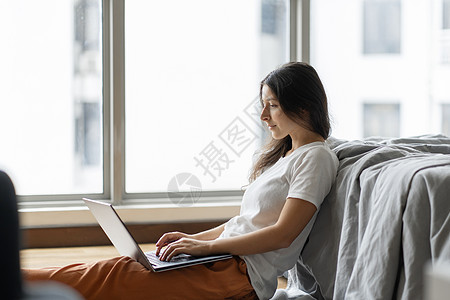 美丽的年轻黑发女孩在笔记本电脑上工作 坐在靠近全景窗户的床边的地板上 时尚现代的内饰 舒适的工作场所 在互联网上购物网络女性地面图片