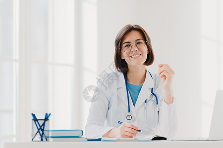 年轻女医生在诊所写纸 为病人开处方 在医院做文书工作 穿着白色医用长袍 坐在舒适的现代办公室里 人 职业 工作概念图片