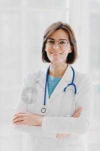 美丽开朗的女医生穿着白色长袍 眼镜和语音内窥镜 双手交叉 自信地看着镜头 留着黑色短发 站在白色背景下 在诊所工作图片