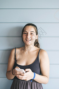 美丽的年轻女士在使用智能手机时靠着木墙上裙子技术成人休闲装女性马尾辫衣服微笑快乐幸福图片