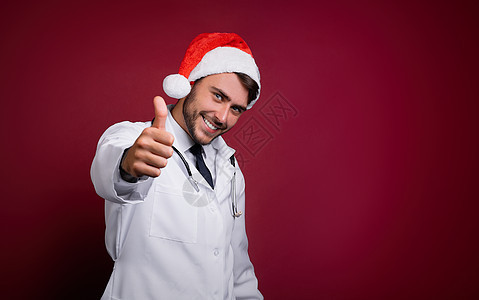 身穿白制服和圣诞老人帽子的英俊年轻医生 站在工作室里 用红色背景微笑和手指在摄像机中横幅新年保健假期男性医院手势诊所医师药品图片