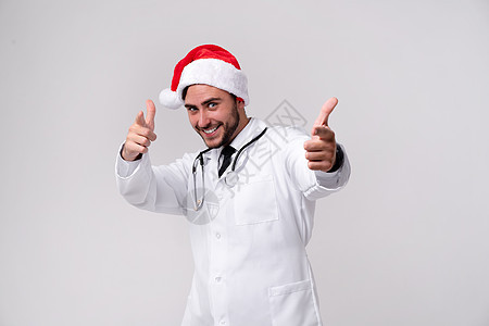 身穿白制服和圣诞老人帽子的英俊年轻医生 站在工作室中 用白色背景微笑和手指进行照相学生专家假期幸福卫生中年诊所男人职业庆典图片