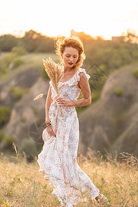 穿着白外衣的美丽年轻女孩 在日落时走在田地里 带了根花束太阳场地村庄稻草裙子小麦微笑女性收成植物图片