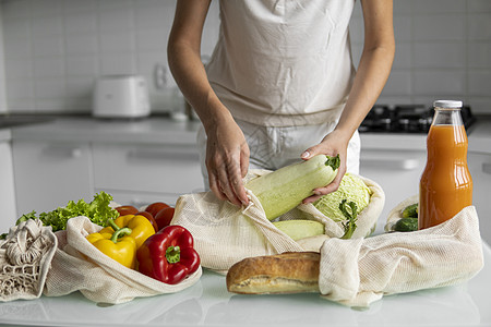 女人的手 拿着一个可重复使用的食品袋 在家里的厨房里放着蔬菜 把西葫芦拿出来 零废物和无塑料概念 蔬菜网眼棉质购物包 生态回收农图片