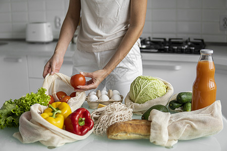 女人的手 拿着一个可重复使用的食品袋 在家里的厨房里放着蔬菜 把西红柿拿出来 零废物和无塑料概念 蔬菜网眼棉质购物包 生态食物杂图片