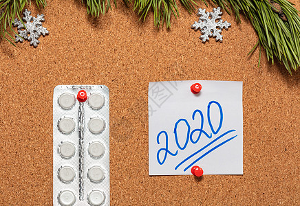 白色药丸 带有 2020 年的白色便签贴在布告板上 布告板上顶部装饰着松树枝和雪花 医疗保健 圣诞节 新年庆典 新常态概念背景图片