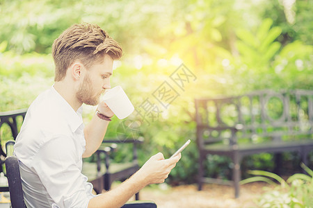 商业英俊的年轻帅哥 喝咖啡 用手机Pho办公室花园咖啡店工具公园男人杯子技术工作微笑图片
